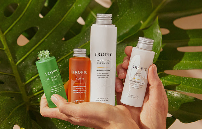 A pair of hands holds four Tropic skincare products against a backdrop of large, green tropical leaves. The products include Rainforest Dew hydration serum, Elixir radiance-boosting omega oil,  Smoothing Cleanser, and Skin Feast moisturiser, all labelled as certified organic. The scene is fresh and vibrant, emphasising natural beauty and skincare.